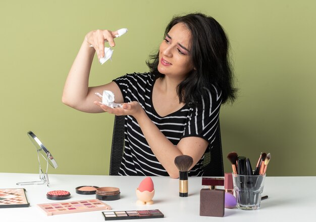 Beeindrucktes junges, schönes Mädchen sitzt am Tisch mit Make-up-Tools, die Haarcreme isoliert auf olivgrüner Wand halten und betrachten