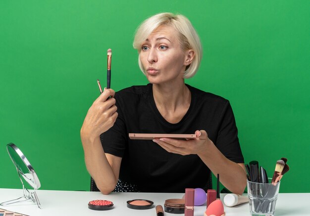 Beeindrucktes junges schönes Mädchen sitzt am Tisch mit Make-up-Tools, die Lidschatten mit Make-up-Pinsel auf grüner Wand auftragen