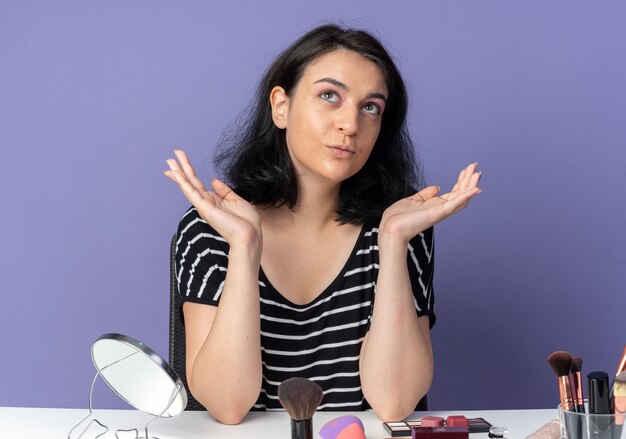 Beeindrucktes junges schönes Mädchen sitzt am Tisch mit Make-up-Werkzeugen, die die Hände einzeln auf der blauen Wand ausbreiten