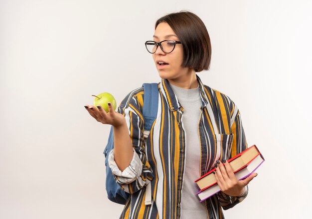 Beeindrucktes junges Studentenmädchen, das Brille und Rückentasche hält Bücher und Apfel betrachtet Apfel lokalisiert auf Weiß