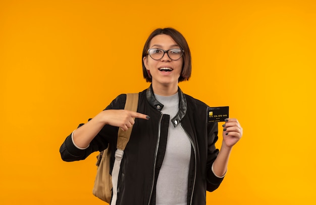 Beeindrucktes junges Studentenmädchen, das Brille und Rückentasche hält und auf Kreditkarte lokalisiert auf Orange zeigt