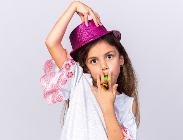 Beeindrucktes kleines kaukasisches Mädchen mit lila Partyhut, der Partypfeife bläst und die Hand auf den Hut legt, isoliert auf weißer Wand mit Kopierraum