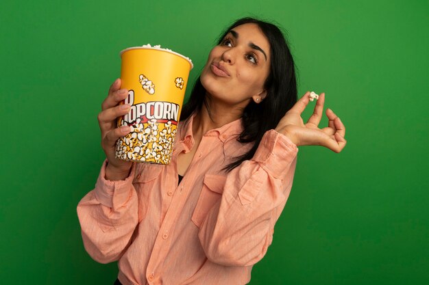 Beeindrucktes Nachschlagen des jungen schönen Mädchens, das rosa T-Shirt hält, das Eimer Popcorn und Popcornfrieden hält, lokalisiert auf grüner Wand