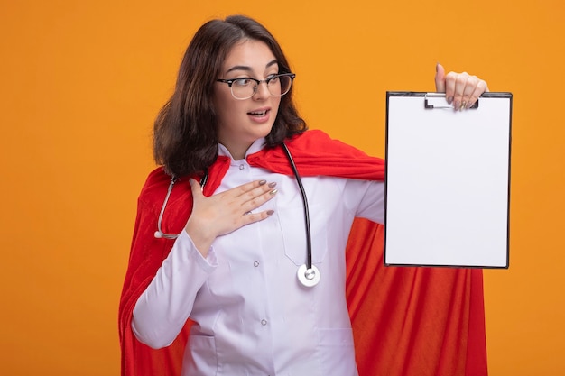 Beeindrucktes Superheldenmädchen in rotem Umhang in Arztuniform und Stethoskop mit Brille, die die Zwischenablage nach vorne zeigt und die Hand auf die Brust legt