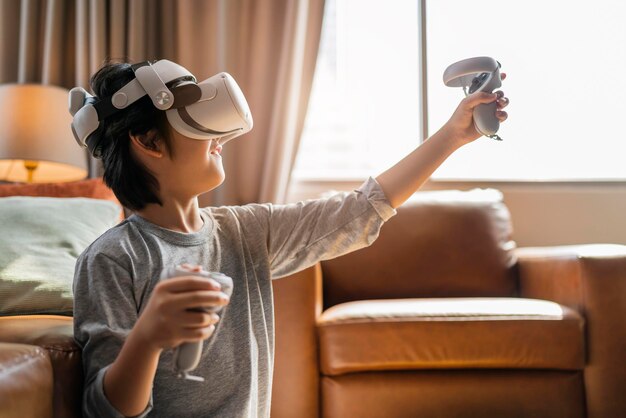 Beendendes asiatisches Kind, männlicher Junge, genießt Metaverse-Gaming mit tragbarem vr-Headset mit Steuergriff, das Sportspiele online im Wohnzimmer zu Hause spielt, junger Teenager mit vr-Technologie
