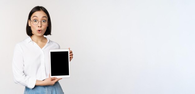 Begeisterte asiatische Büroangestellte mit Brille, die einen digitalen Tablet-Bildschirm zeigt, der Informationen auf dem Gadget-Display zeigt, das über weißem Hintergrund steht