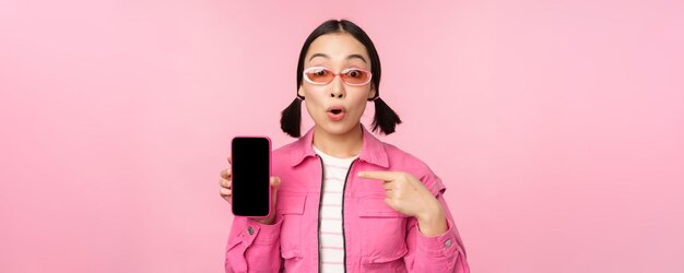 Begeisterte asiatische Frau in stylischer Kleidung, Sonnenbrille, die mit dem Finger auf den Bildschirm des Mobiltelefons zeigt und die Smartphone-Anwendung zeigt, die über rosa Hintergrund steht