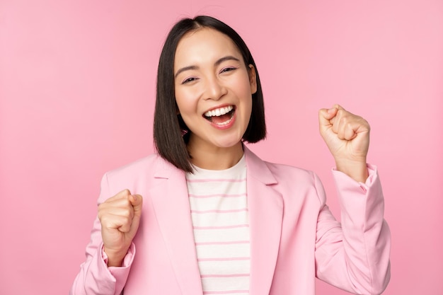 Begeisterte asiatische geschäftsfrau der verkäuferin sagt ja, erreicht das ziel und feiert den triumphalen blick mit freude und lächelndem stehen über rosa hintergrund