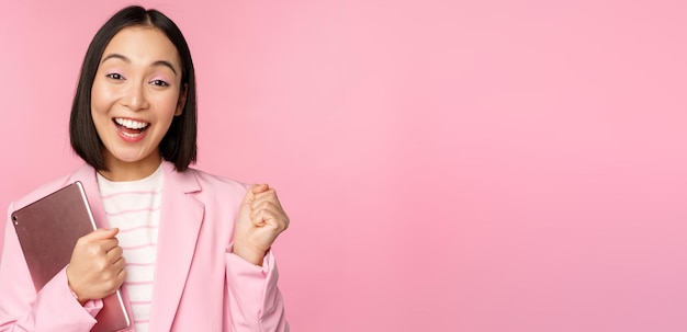 Begeisterte asiatische Geschäftsfrau mit digitalem Tablet, die vor Freude schreit und über rosa Hintergrund im Anzug triumphiert Kopierraum