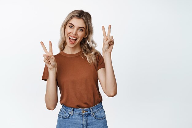 Begeisterte blonde Frau zeigt Friedenszeichen und lächelt lachend positiv über weißem Hintergrund