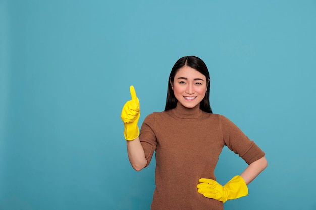 Begeisterte, fröhliche asiatische Hausarbeiterin mit Daumen nach oben von der Hausarbeit und mit gelben Handschuhen für die Handsicherheit, Reinigungskonzept, glückliches lächelndes Hausmädchen mit positivem Gemütszustand
