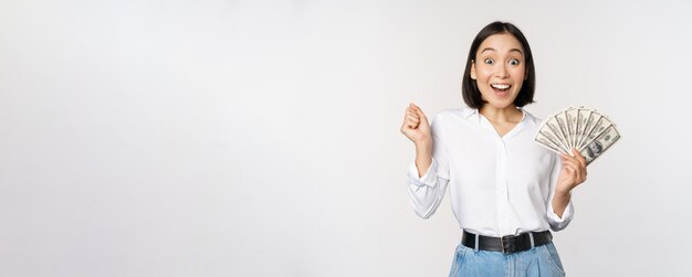 Begeisterte junge asiatische Frau, die aufgeregt in die Kamera schaut, die Gelddollar in der Hand hält, die über weißem Hintergrund steht