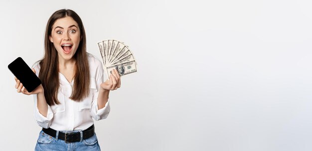 Begeisterte junge Frau gewinnt Geld mit Smartphone-App-Schnittstelle und Bargeld-Mikrokreditpreiskonzept, das über weißem Hintergrund steht