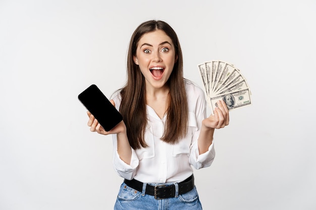 Begeisterte junge Frau gewinnt Geld, zeigt Smartphone-App-Schnittstelle und Bargeld, Mikrokredit, Preiskonzept, steht auf weißem Hintergrund.