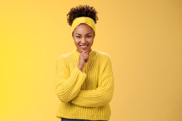 Begeisterte, kreative, gut aussehende schwarze junge Frau mit Pullover-Stirnband, ausgezeichneter Plan, lächeln, zufrieden, begeistert aussehen, nachdenkliche Kamera-Make-up-Idee, gelber Hintergrund.