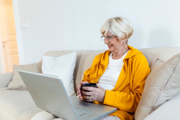 Begeisterte progressive Seniorin winkt mit der Hand und fühlt sich glücklich, während sie einen Videoanruf auf dem Laptop nutzt und mit Enkelkindern spricht Ältere Frau, die auf dem Sofa sitzt und eine Tasse Kaffee hält