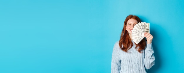 Kostenloses Foto begeisterte rothaarige frau, die geld in der nähe des lächelnden gesichtes zeigt, das zufrieden steht, bereit zum einkaufen über b