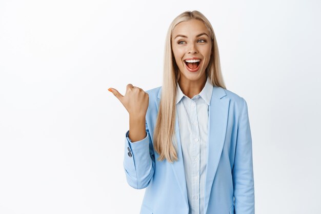 Begeisterte Verkäuferin, die mit dem Finger nach links zeigt und erstaunt auf den Verkauf des Logos der Firma blickt, der im Anzug vor weißem Hintergrund steht