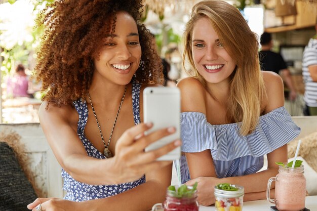 Begeisterte weibliche Models gemischter Rassen haben Spaß zusammen, posieren für Selfies auf Smartphones, lächeln breit und lächeln im Café mit Smoothie und Cocktails. Menschen, ethnische Zugehörigkeit und Freizeit