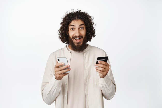 Begeisterter arabischer Typ, der Handy und Kreditkarte hält, lächelt erstaunt über den weißen Hintergrund der Kamera
