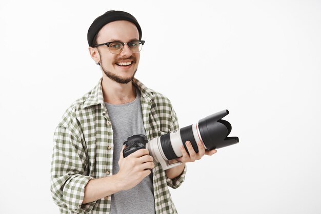 begeisterter gut aussehender reifer Mann in Brille und schwarzer Mütze, die professionelle Kamera hält und vor Freude lächelnd als Journalist oder Fotograf lächelt