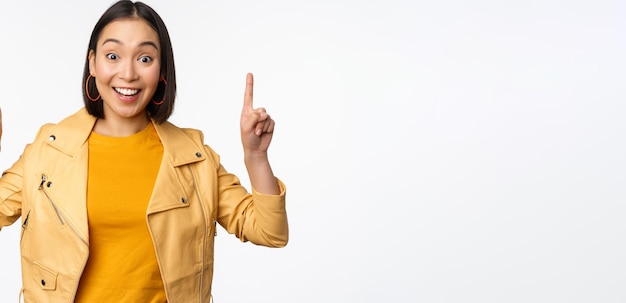 Begeistertes asiatisches Mädchen, das mit dem Finger nach oben zeigt und Werbung auf der Oberseite zeigt, die glücklich lächelt und das Promo-Angebot oder Banner vor weißem Hintergrund demonstriert