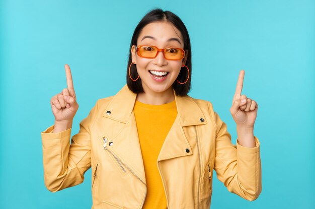 Begeistertes asiatisches Mädchen mit Sonnenbrille zeigt mit den Fingern nach oben und zeigt Banner oder Logo auf den oberen Ständern auf blauem Hintergrund