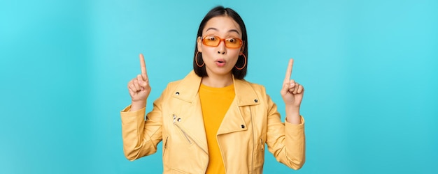 Begeistertes asiatisches Mädchen mit Sonnenbrille zeigt mit den Fingern nach oben und zeigt Banner oder Logo auf der Oberseite steht über blauem Hintergrund Kopierbereich