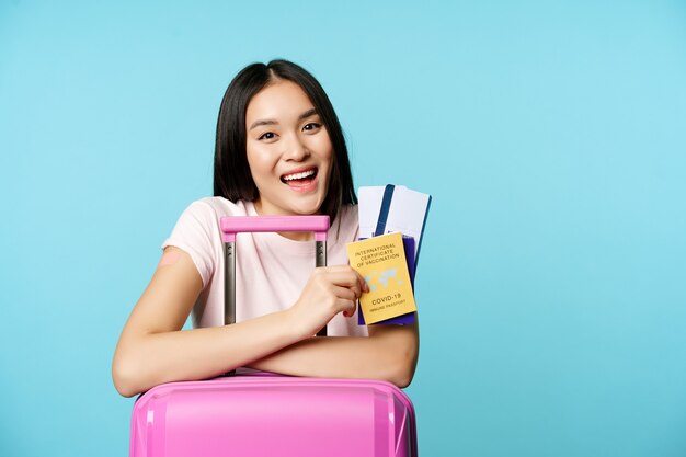 Begeistertes asiatisches Mädchen zeigt Passtickets und Gesundheitskovid-Impfzertifikat lächelnd...