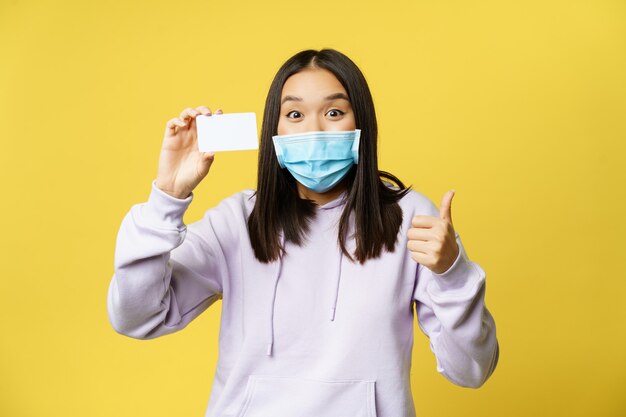 Begeistertes koreanisches Mädchen in Gesichtsmaske, Karte und Daumen nach oben zeigend, Produkt empfehlen, einkaufen, auf gelbem Hintergrund stehend.