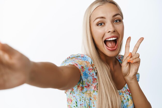 Begeistertes schönes blondes Mädchen, das selfie mit dem Friedenszeichen macht und glücklich stehend über weißem Hintergrund lächelt