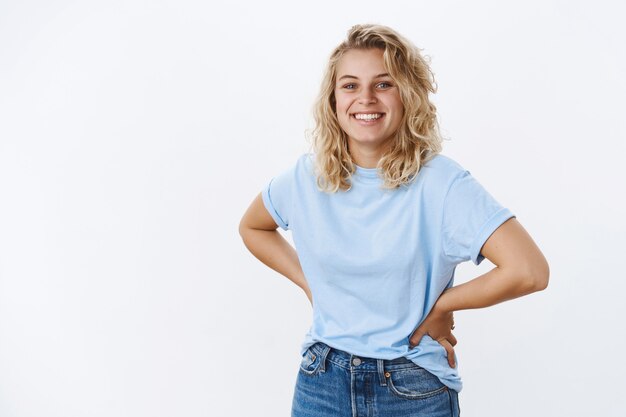 Begeistertes und fröhliches, süßes, lächelndes blondes Mädchen mit lockiger Frisur, das in entspannter und glücklicher Pose die Hände an der Taille hält, während es sorglos grinst und gute Laune und Spaß über grauer Wand hat