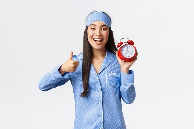 Begeistertes und glücklich lächelndes asiatisches Mädchen in blauem Pyjama und Schlafmaske, das Wecker und Daumen hoch zur Zustimmung zeigt, wie das frühe Aufwachen für den Morgenlauf, einen aktiven und gesunden Lebensstil