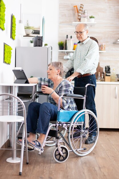 Behinderte ältere Frau im Rollstuhl mit Tablet-Computer in der Küche mit Ehemann in der Nähe. Gelähmte behinderte alte ältere Person, die moderne Online-Internet-Web-Technologie verwendet.