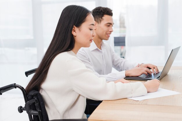 Behinderte asiatische junge Frau, die auf dem Rollstuhl arbeitet mit seinem Kollegen verwendet Laptop auf Tabelle im Büro sitzt