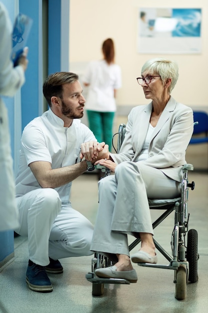 Behinderte, reife Frau im Rollstuhl, die mit einem männlichen Arzt spricht, während sie im Krankenhausflur Händchen hält