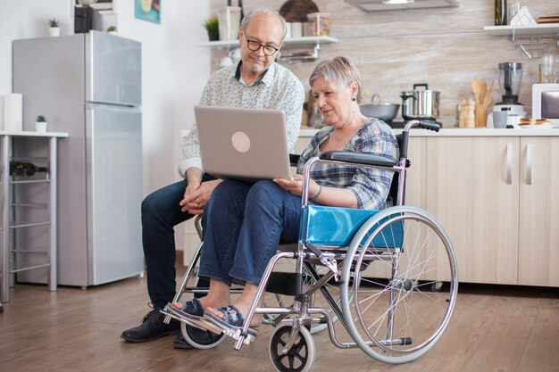 Behinderte Seniorin im Rollstuhl und ihr Ehemann haben eine Videokonferenz auf einem Tablet-PC in der Küche. Gelähmte alte Frau und ihr Mann bei einer Online-Konferenz.