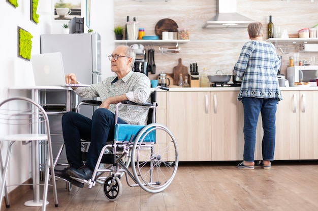 Behinderter älterer Mann im Rollstuhl, der von zu Hause aus am Laptop in der Küche arbeitet, während die Frau das Frühstück kocht. Behinderter Geschäftsmann, Behinderten-Unternehmerlähmung für ältere Menschen im Ruhestand.