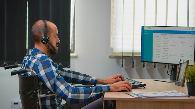 Behinderter behinderter Operator, der im Rollstuhl sitzt und Telemarketing im Geschäftsbüro macht. Immobilisierter, behinderter, gelähmter Freiberufler, der in einem Finanzunternehmensgebäude mit Heatset arbeitet