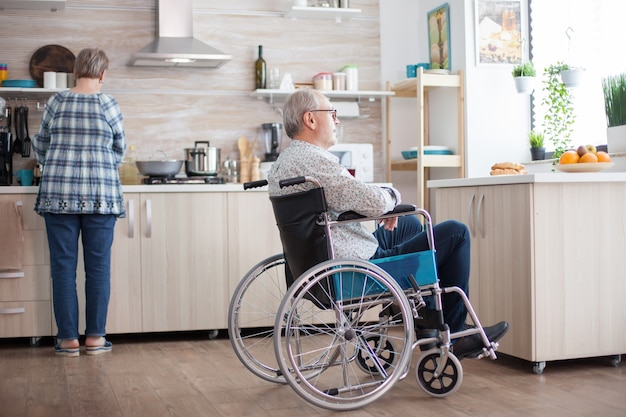 Behinderter Mann sitzt im Rollstuhl in der Küche und schaut durch das Fenster, während die Frau das Frühstück zubereitet. Invalide, Rentner, Behinderte, Lähmung.