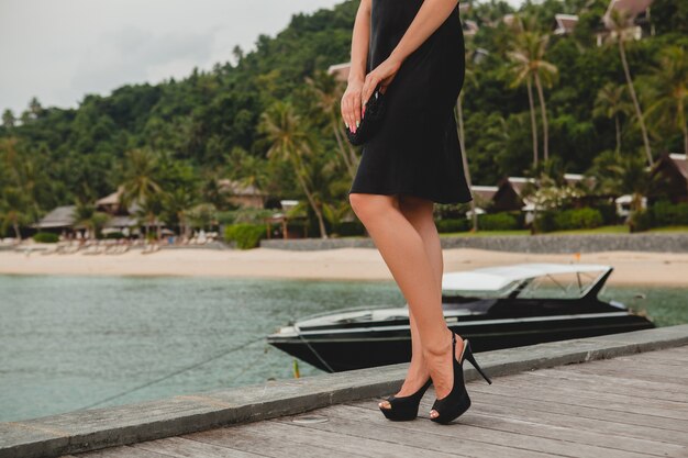 Beine in hochhackigen schwarzen Schuhen der sexy sexy attraktiven Luxusfrau gekleidet im schwarzen Kleid, das auf Pier im Luxusresorthotel, Sommerferien, tropischer Strand aufwirft