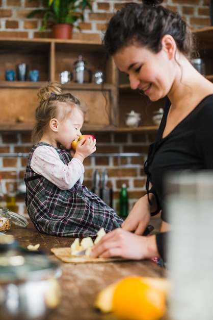 Beißender Apfel des Mädchens nahe, Mutter zu kochen