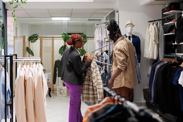 Bekleidungsgeschäftsassistent hilft Kunden bei der Auswahl des Hemdes in der Bekleidungsabteilung des Einkaufszentrums. Modeboutique afroamerikanische Frau, die dem Kunden trendiges Kleidungsstück zeigt