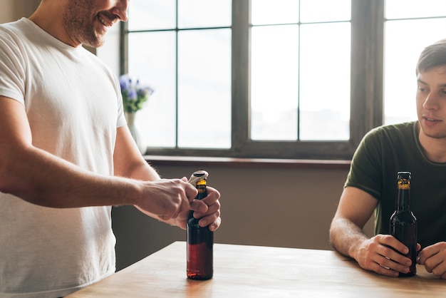 Bemannen Sie das Betrachten seines Freundes, der die Bierflasche mit Öffner auf Tabelle öffnet