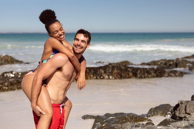 Bemannen Sie das Geben der Frau auf Strand im Sonnenschein huckepack