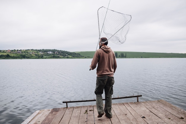 Kostenloses Foto bemannen sie das halten des fischernetzes und der stange, die auf hölzernem pier vor see stehen