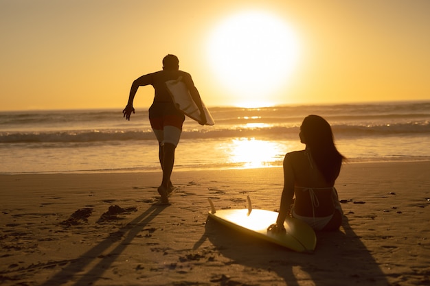 Bemannen Sie das Laufen mit Surfbrett während die Frau, die auf dem Strand während des Sonnenuntergangs sich entspannt