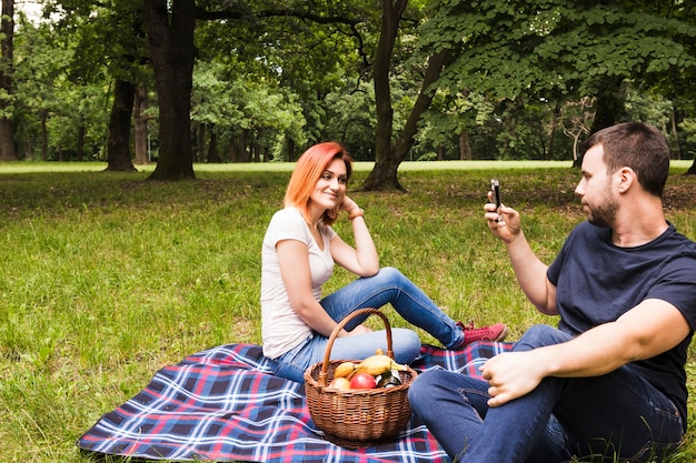 Bemannen Sie das Machen der Fotografie ihrer lächelnden Freundin auf Smartphone am Picknick