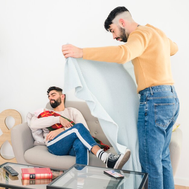 Bemannen Sie das Setzen der Decke über seinem Freund, der zu Hause mit Baby auf Sofa schläft