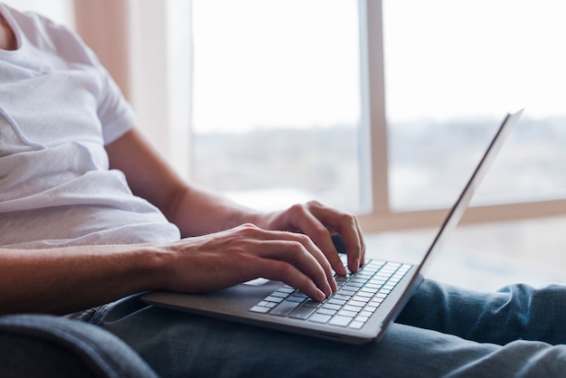 Bemannen Sie das Sitzen auf Stuhl und auf Laptop nahe Fenster schreiben